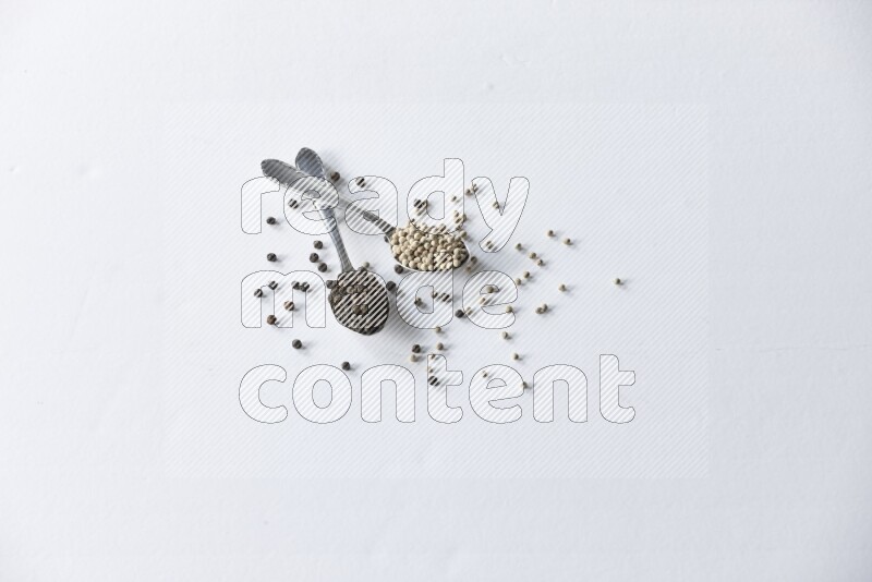 ملعقتان معدنيتان ممتلئتان بحبوب الفلفل الأسود والأبيض مع حبوب متناثرة على أرضية بيضاء