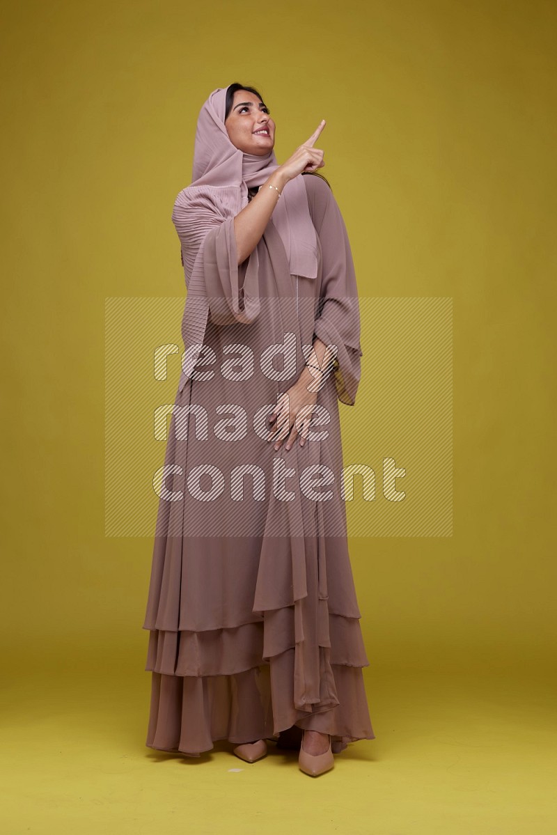 A woman Pointing on a Yellow Background wearing Brown Abaya with Hijab