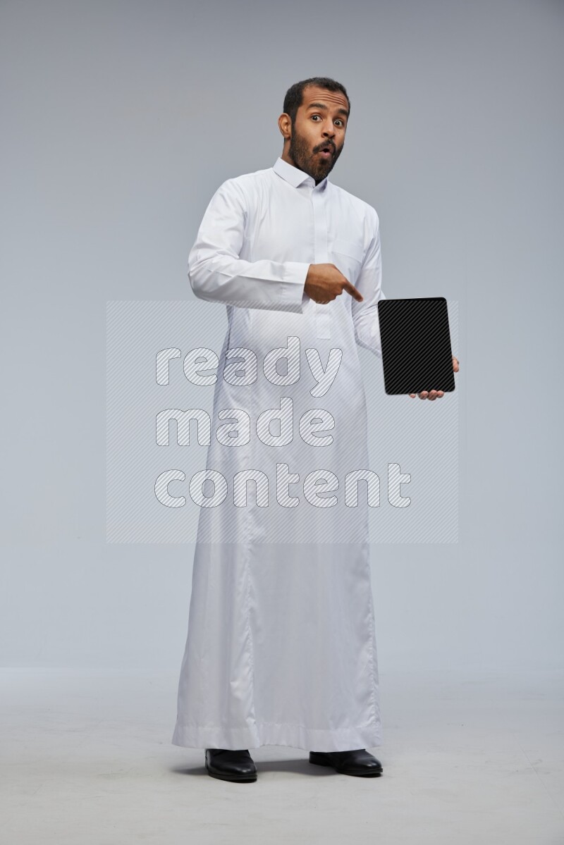 Saudi man wearing Thob standing showing tablet to camera on Gray background