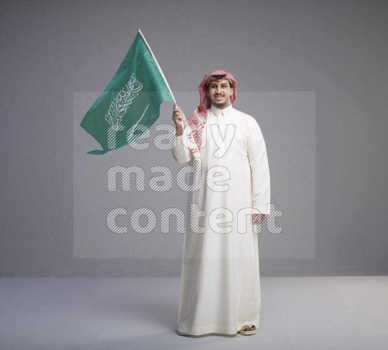 A Saudi man standing wearing thob and red shomag raising big Saudi flag on gray background