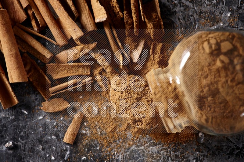 Herbal glass jar full cinnamon powder flipped and a metal spoon full of powder surrounded by cinnamon sticks on textured black background in different angles