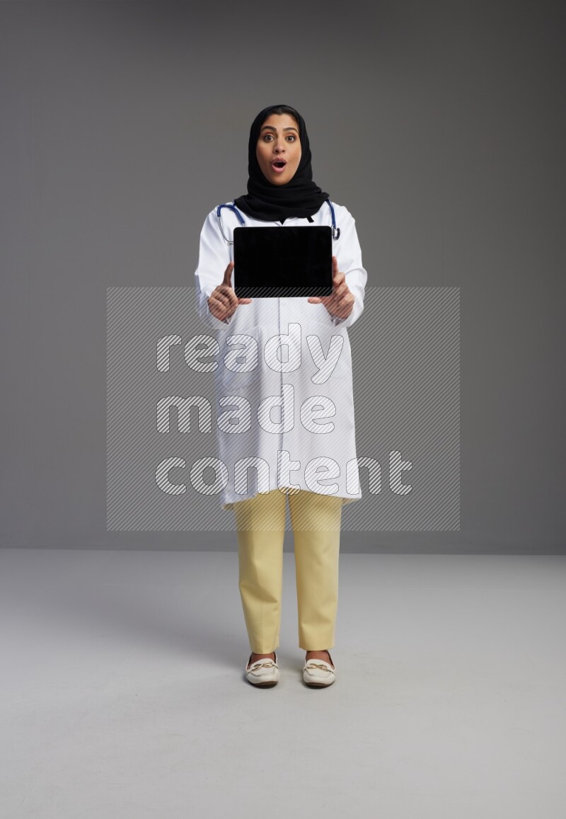 Saudi woman wearing lab coat with stethoscope standing showing tablet to camera with sign in the back on Gray background