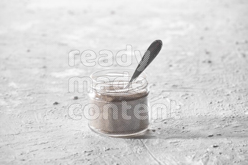 A glass jar full of black pepper powder and a metal spoon full of powder on a textured white flooring