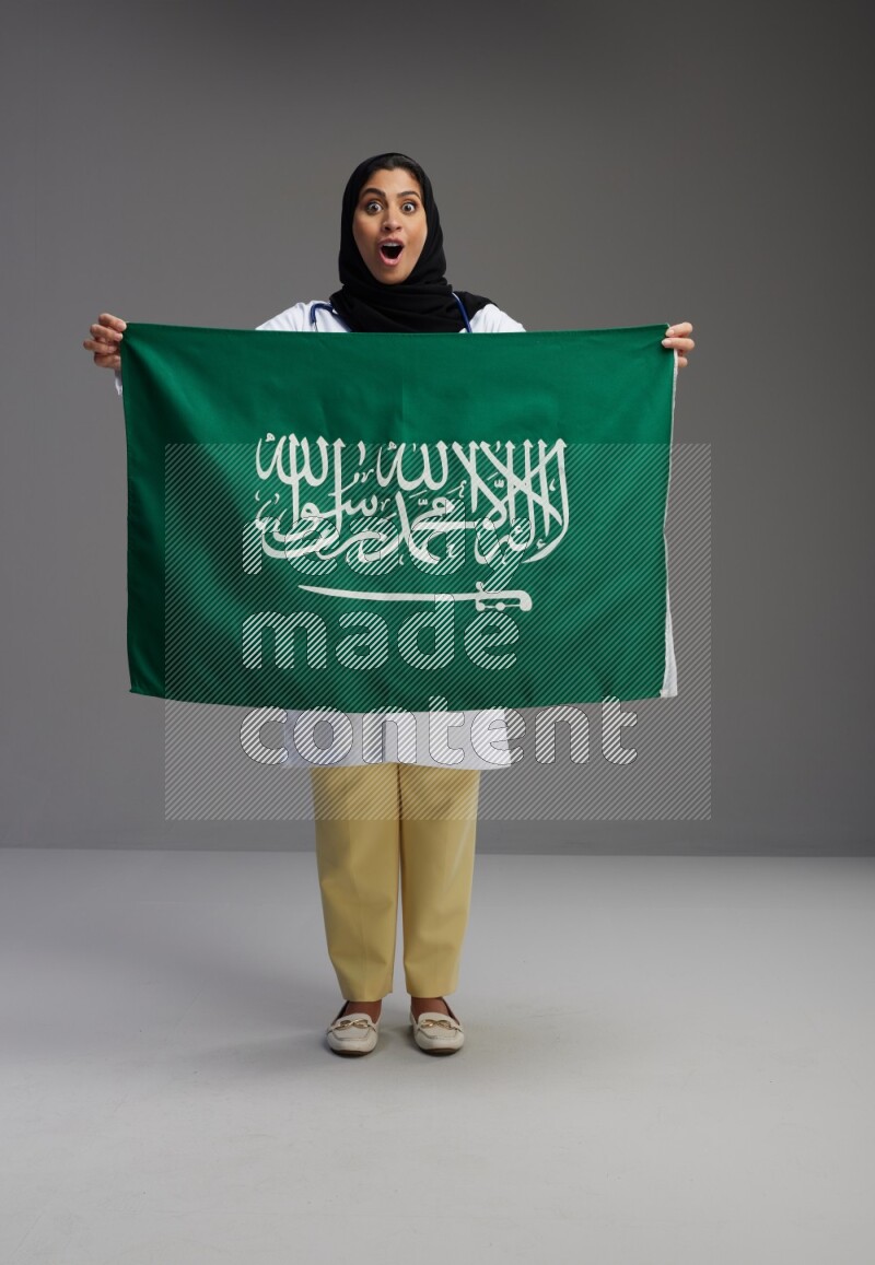 Saudi woman wearing lab coat with stethoscope standing holding Saudi flag on Gray background