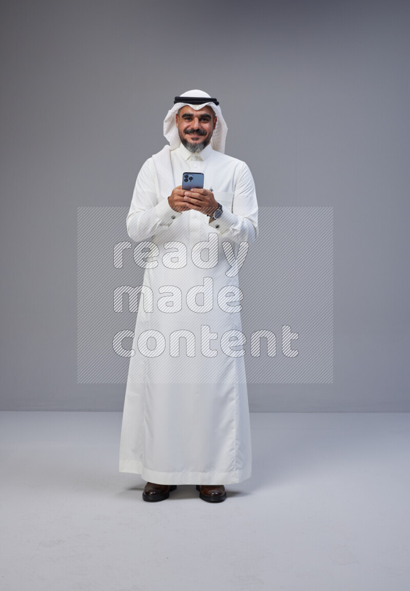 Saudi man Wearing Thob and white Shomag standing texting on phone on Gray background