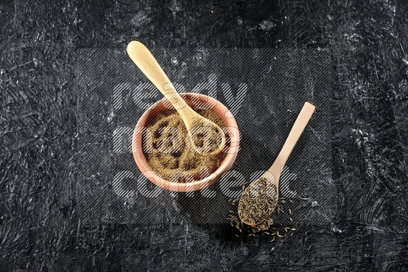 A wooden bowl and 2 wooden spoons full of cumin powder and cumin seeds on a textured black flooring