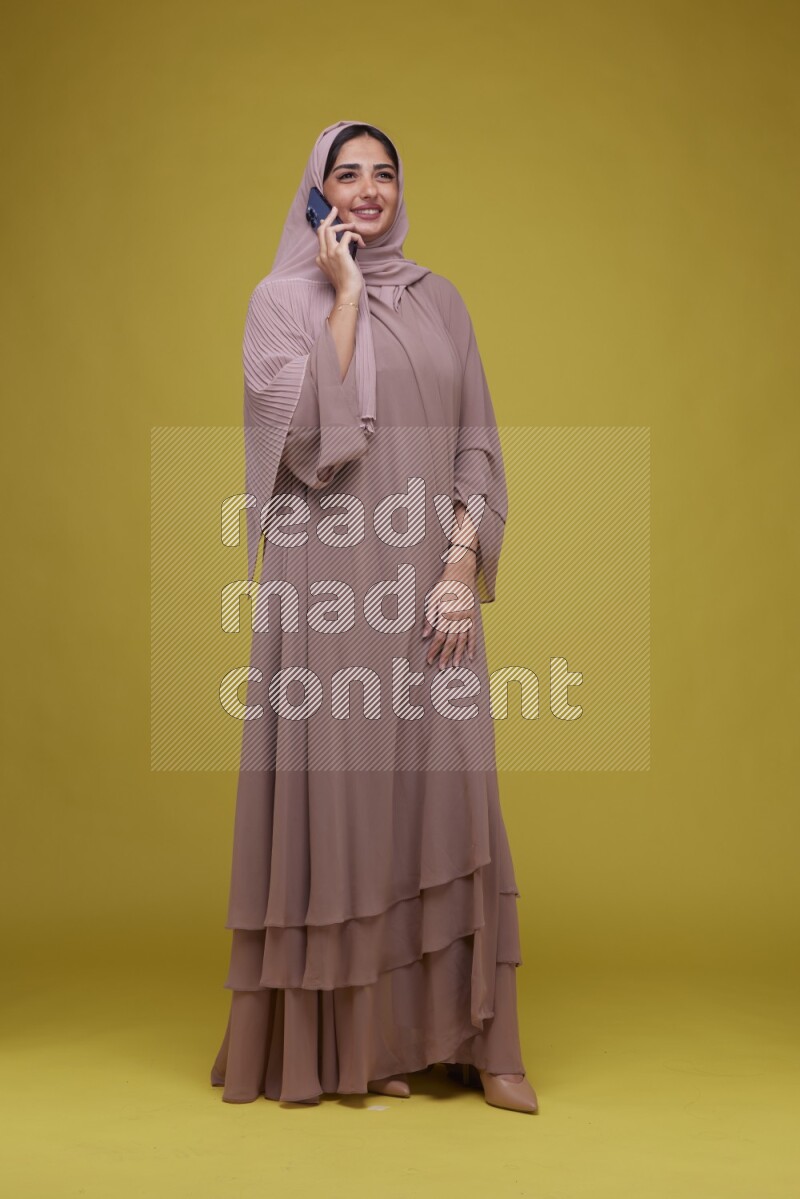 A woman having a call on a Yellow Background wearing Brown Abaya with Hijab