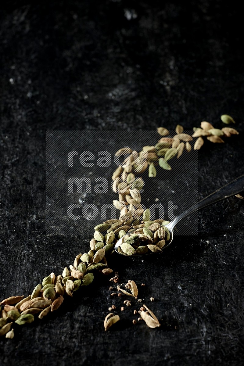 A Metal spoon full of cardamom seeds and some seeds beside it on a textured black flooring