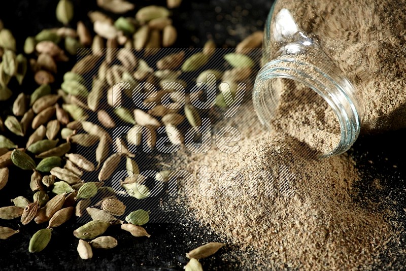 A flipped glass spice jar full of cardamom powder and powder spilled out of it with cardamom seeds spreaded on textured black flooring