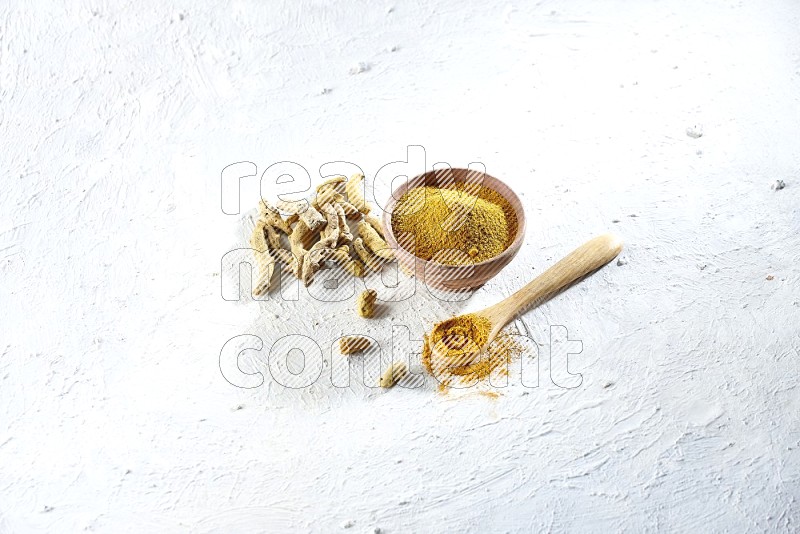 وعاء خشبي وملعقة خشبية ممتلئان ببودرة الكركم وأصابع الكركم المجففة بجانبهما على خلفية بيضاء