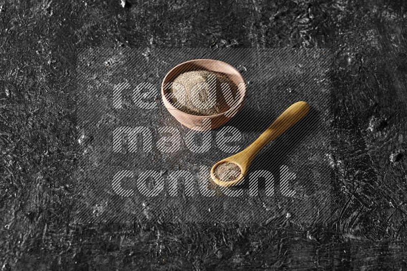 A wooden bowl and wooden spoon full of black pepper powder on a textured black flooring