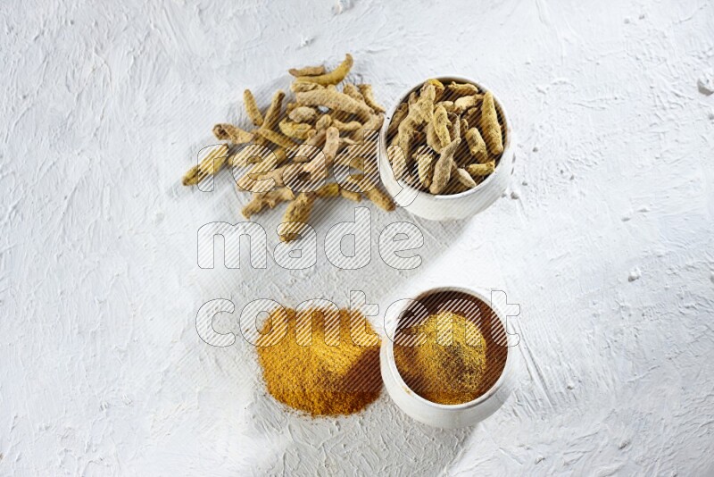 وعاءان فخاريان بيج ممتلئان ببودرة الكركم وأصابع الكركم المجففة، مع بودرة وأصابع الكركم بجانبهما على خلفية بيضاء