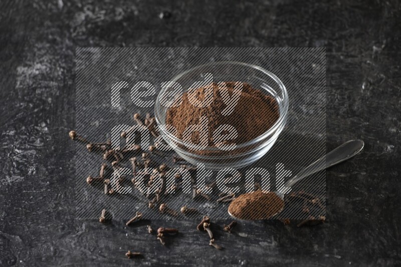 A glass bowl and a metal spoon full of cloves powder with gloves grains beside them on a textured black flooring