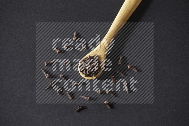 A wooden spoon full of cloves and some spreaded out of it on a black flooring