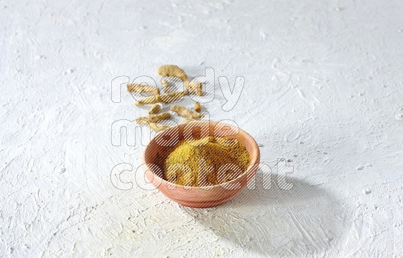 A wooden bowl full of turmeric powder with dried whole fingers on textured white flooring