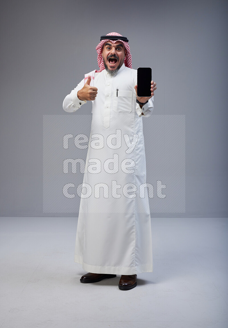 Saudi man Wearing Thob and red Shomag standing showing phone to camera on Gray background