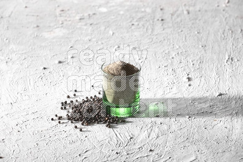 A glass cup full of black pepper powder and black pepper spread on a textured white flooring