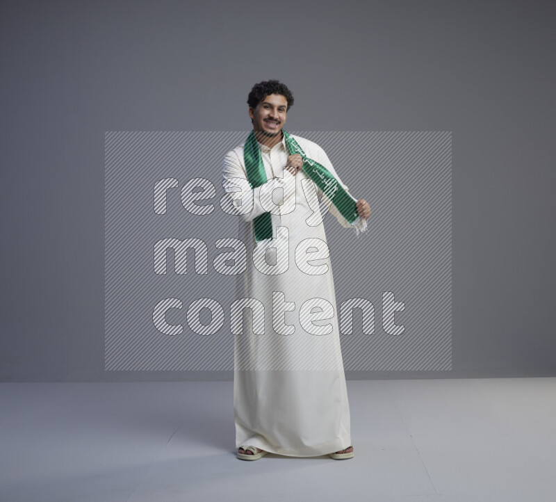 A Saudi man standing wearing thob and Saudi flag scarf on gray background