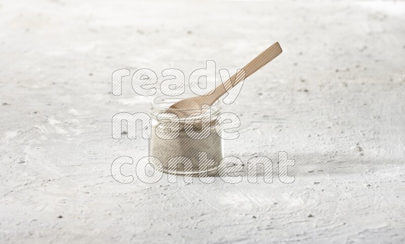 A glass jar and wooden spoon full of white pepper powder on textured white flooring