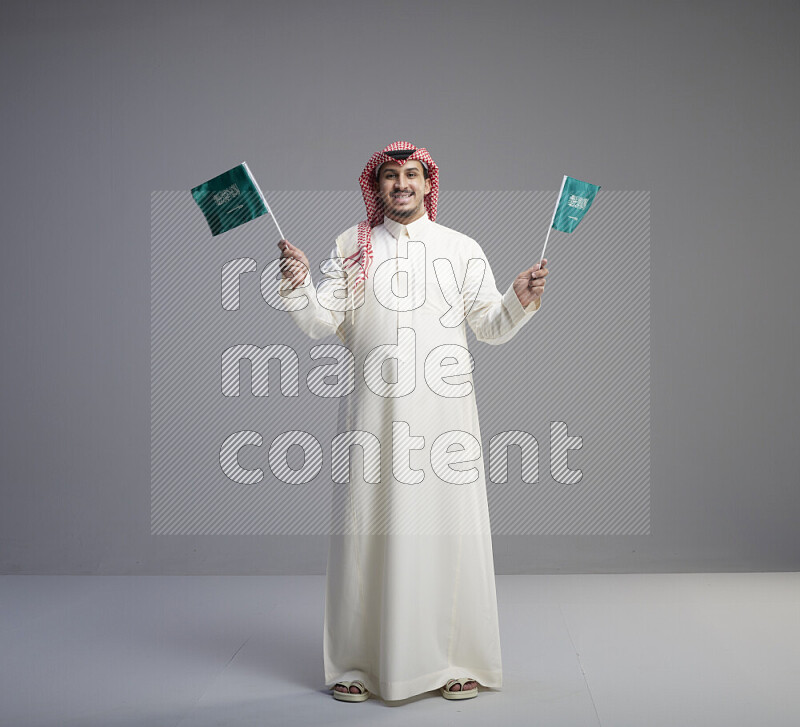 A Saudi man standing wearing thob and red shomag raising small Saudi flag on gray background