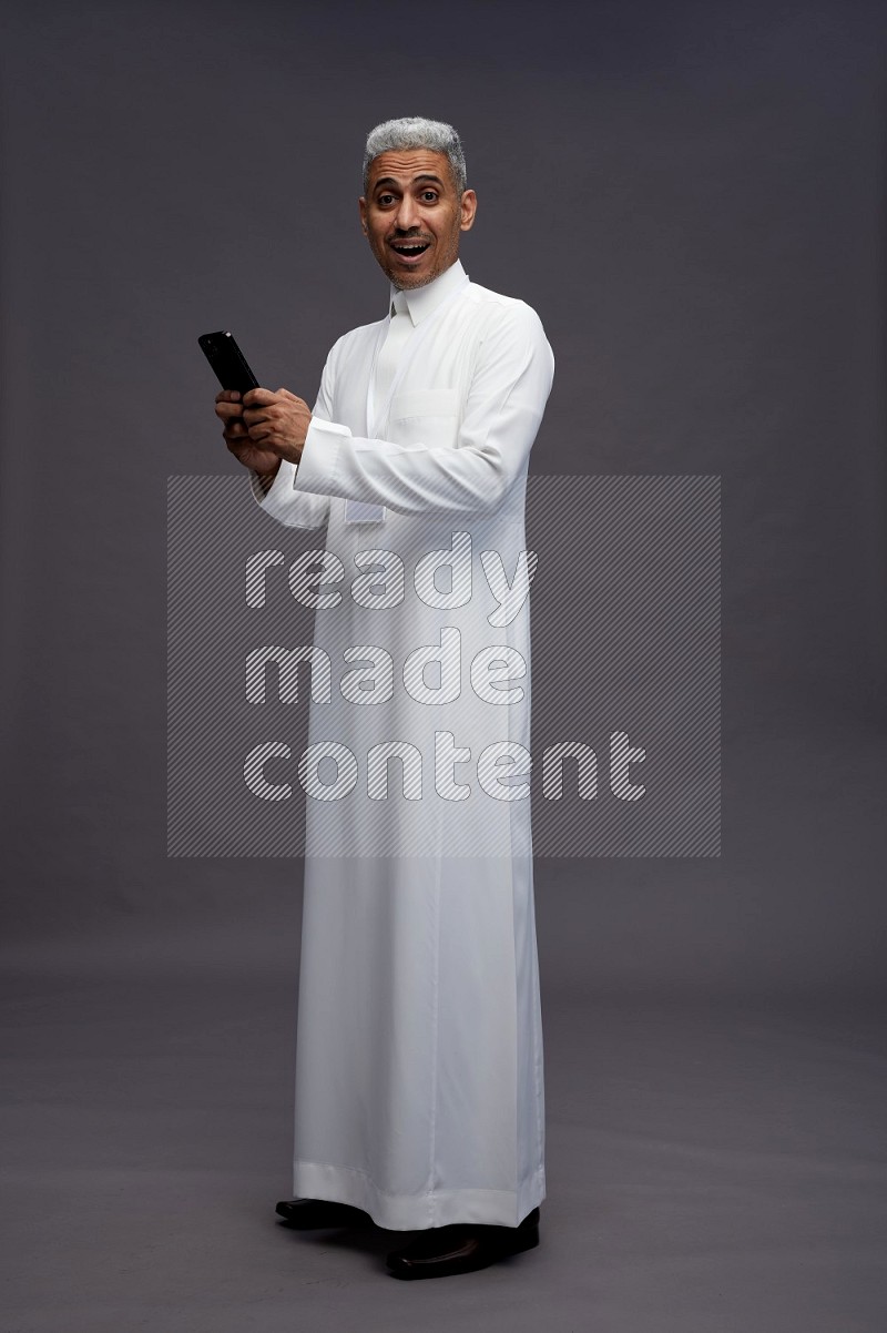 Saudi man wearing thob with neck strap employee badge standing texting on phone on gray background