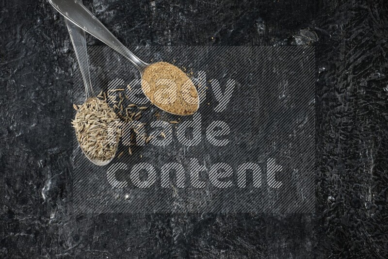 ملعقتان معدنيتان مملوءتان بحبوب الكمون وبودرة الكمون على أرضية سوداء