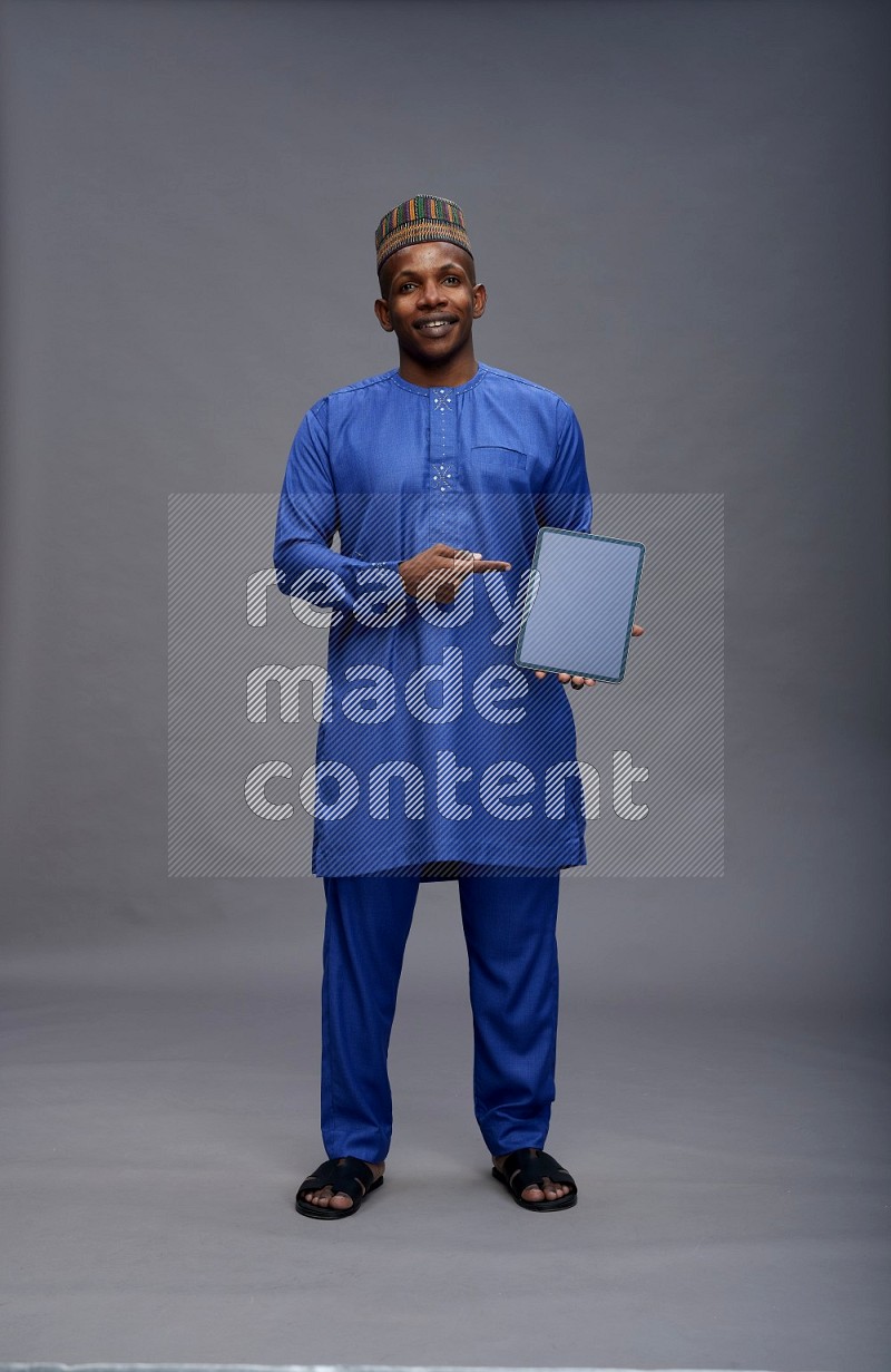 Man wearing Nigerian outfit standing showing tablet to camera on gray background