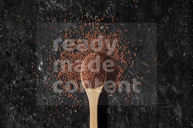 A wooden spoon full of garden cress seeds surrounded by the seeds on a textured black flooring