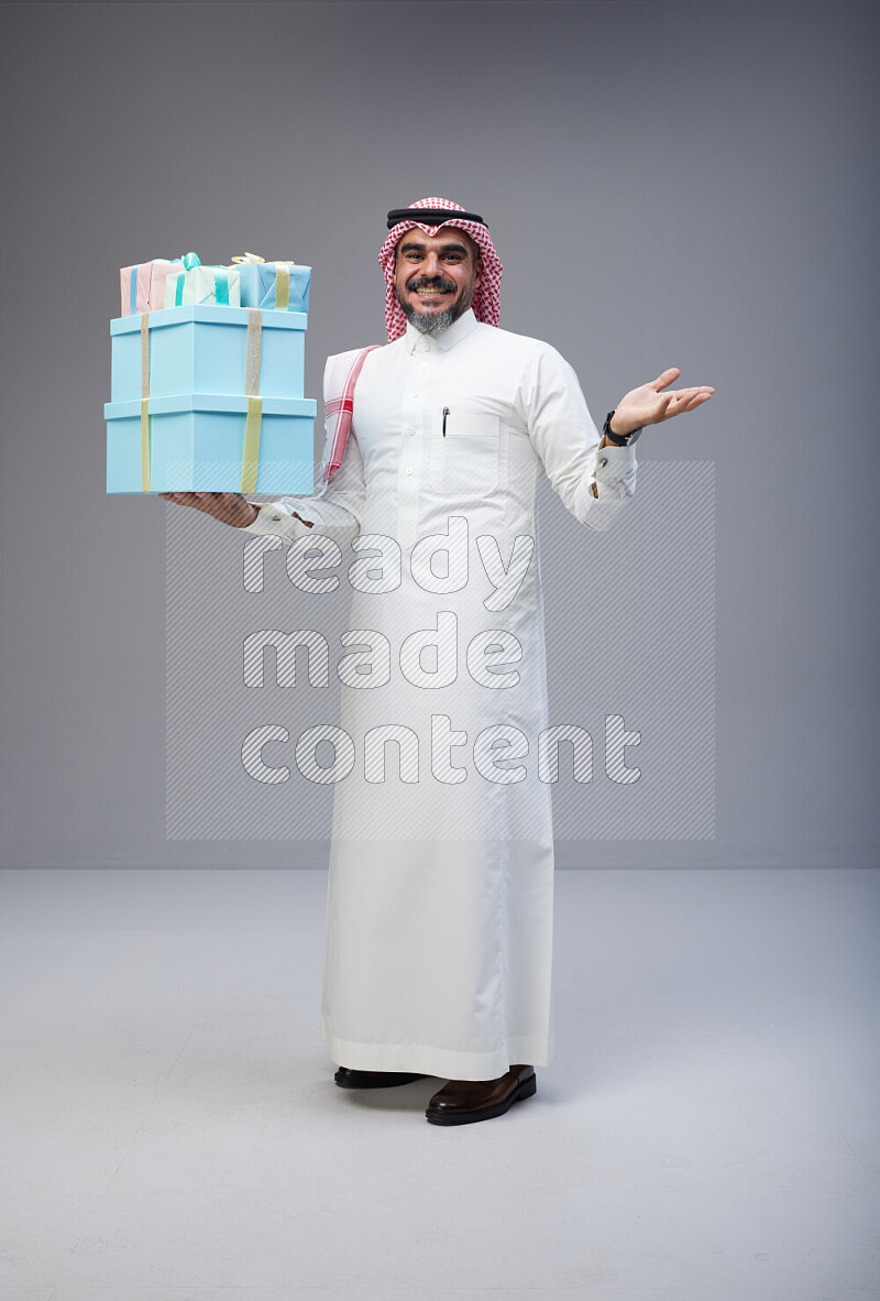 Saudi man Wearing Thob and red Shomag standing holding gift box on Gray background