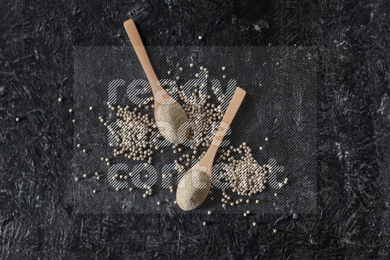 2 wooden spoons full of white pepper powder with white pepper beads on textured black flooring