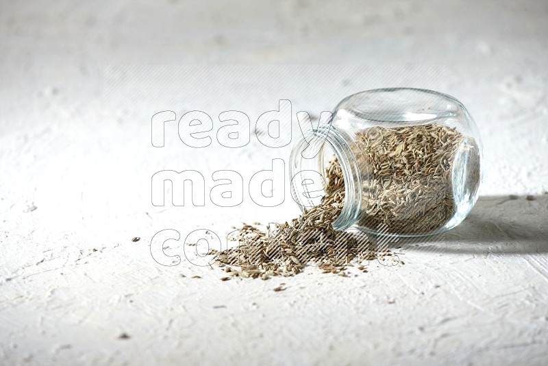 A flipped glass spice jar full of cumin seeds and the seeds spilled out on textured white flooring