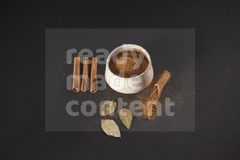 Cinnamon powder in a white pottery bowl and cinnamon sticks and laurel leaves on black background