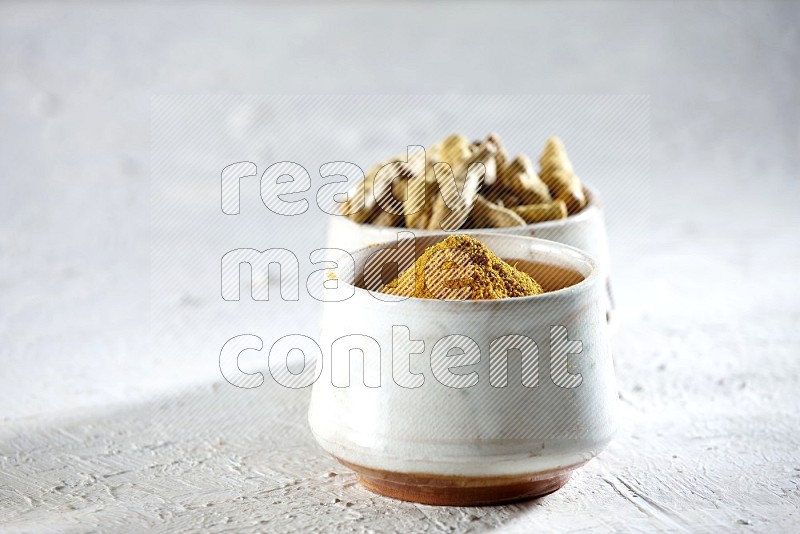 2 beige pottery bowl full of turmeric powder and dried turmeric whole fingers on textured white flooring