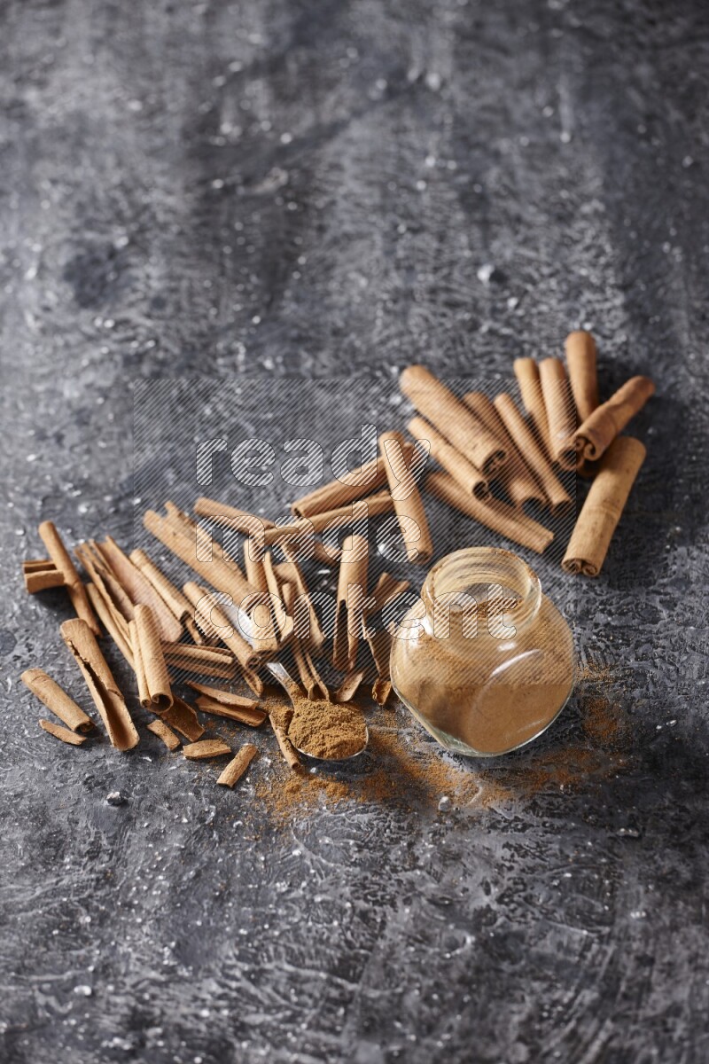 Herbal glass jar and a metal spoon full of cinnamon powder surrounded by cinnamon sticks on textured black background