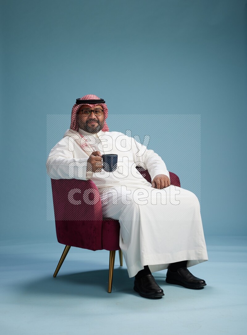 Saudi Man with shimag sitting on chair drinking coffee on blue background
