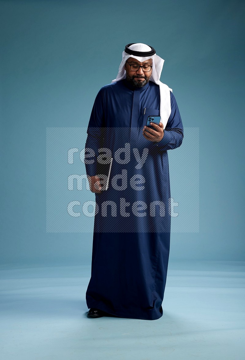 Saudi Man with shimag Standing texting on phone on blue background