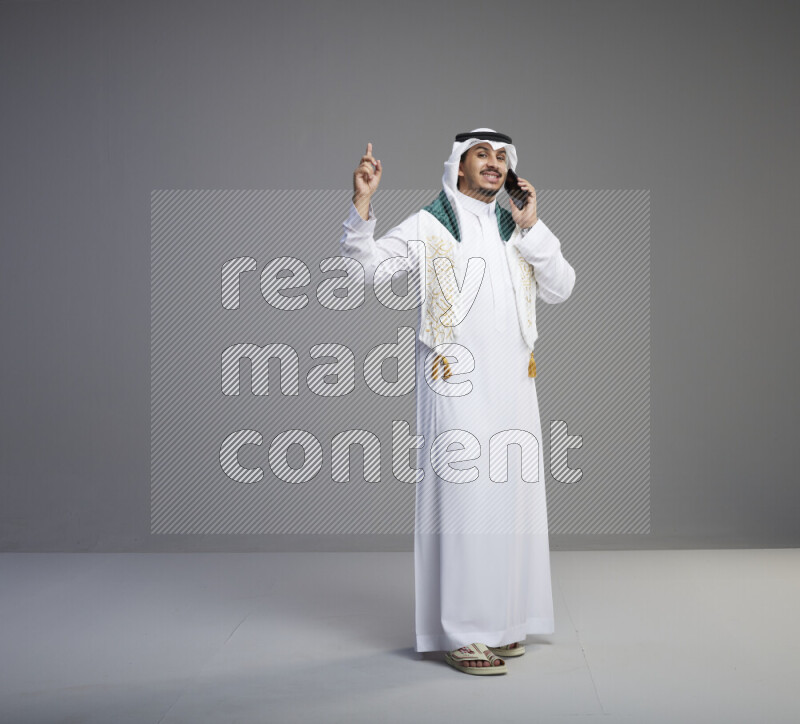 A saudi man standing wearing thob and white shomag with flag scarf on his neck talking on phone on gray background