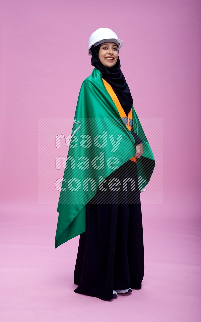 Saudi woman wearing Abaya with engineer vest and helmet standing holding Saudi flag on pink background