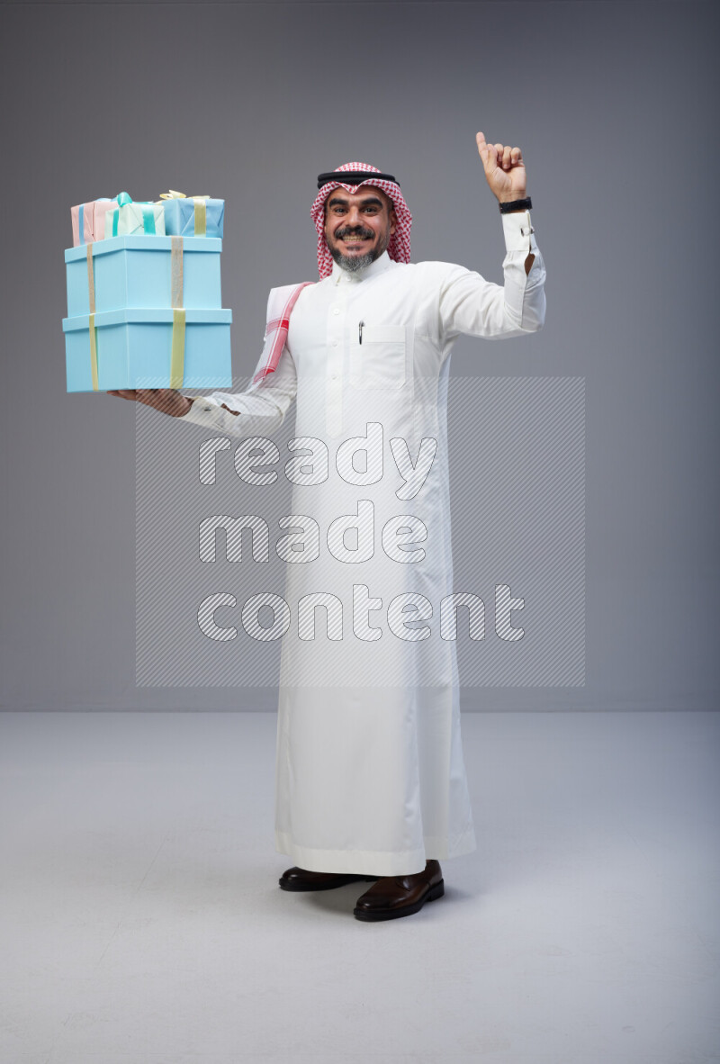 Saudi man Wearing Thob and red Shomag standing holding gift box on Gray background