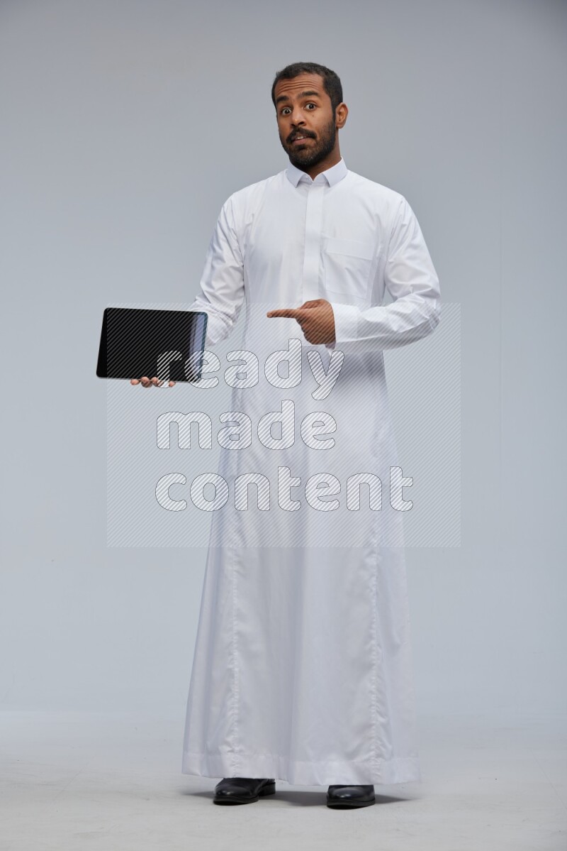 Saudi man wearing Thob standing showing tablet to camera on Gray background