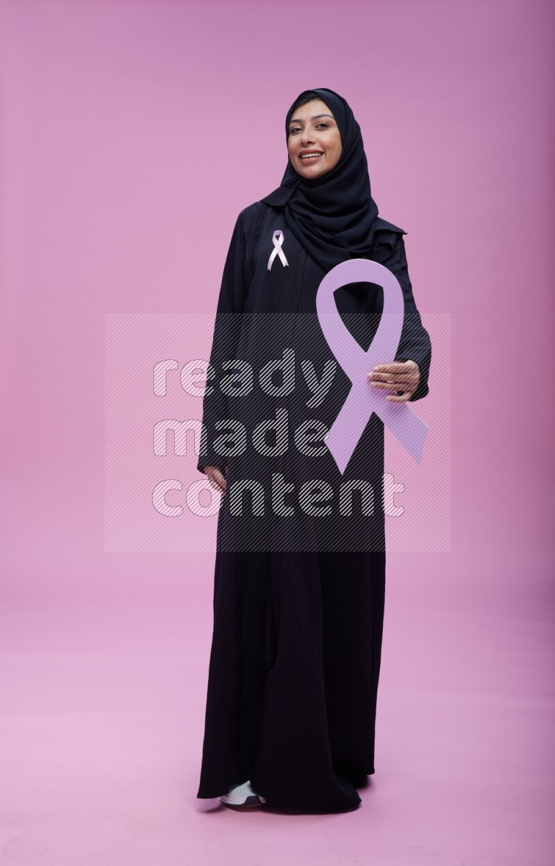 Saudi woman wearing pink ribbon on abaya standing holding awareness ribbon on plain pink background