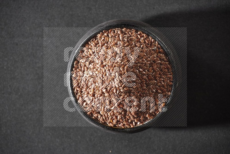 A black pottery bowl full of flaxseeds on a black flooring