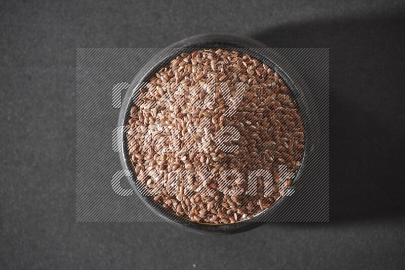 A black pottery bowl full of flaxseeds on a black flooring