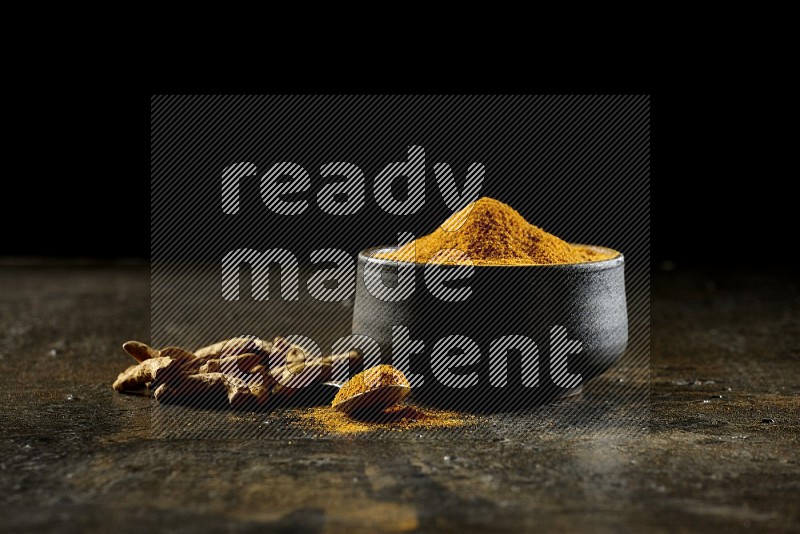 A pottery black bowl and a metal spoon full of turmeric powder with dried turmeric fingers on a textured black flooring