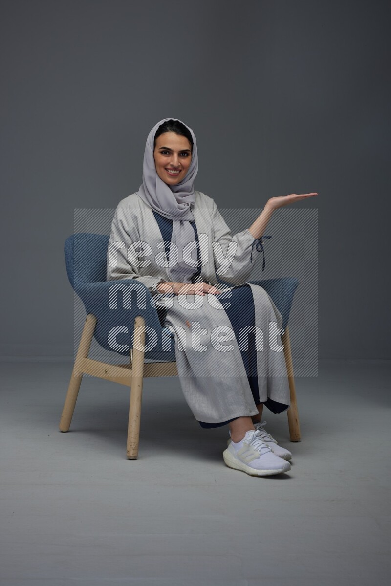 A Saudi woman wearing a light gray Abaya and head scarf sitting on a dark grey chair while making multi hand gestures eye level on a grey background