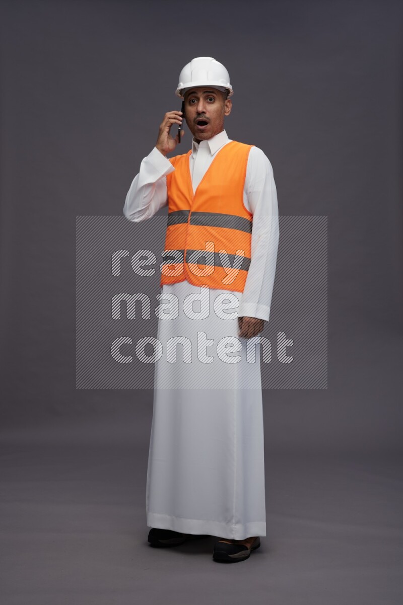 Saudi man wearing thob with engineer vest standing talking on phone on gray background