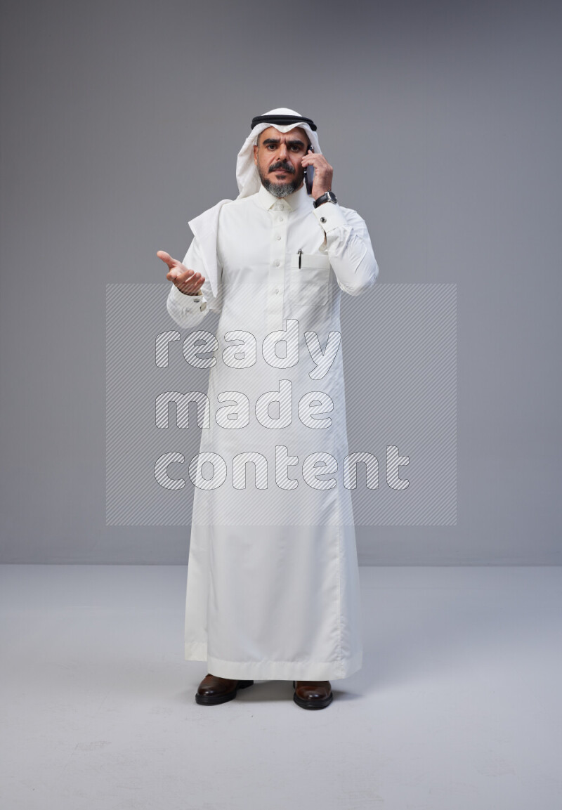 Saudi man Wearing Thob and white Shomag standing talking on phone on Gray background