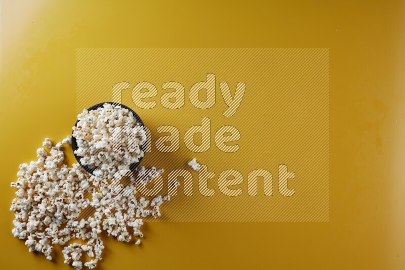A black ceramic bowl full of popcorn with popcorn beside it on a yellow background in different angles