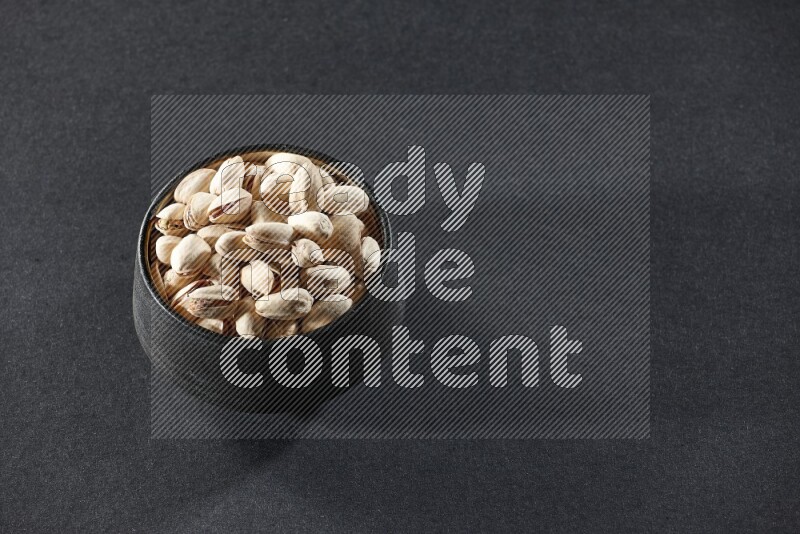 A black pottery bowl full of pistachios on a black background in different angles