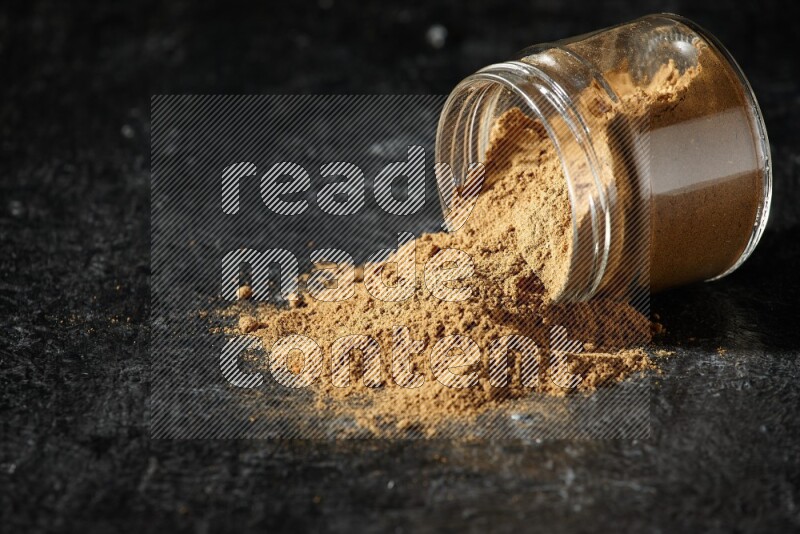 A flipped glass jar full of allspice powder and powder spilled out of it on a textured black flooring